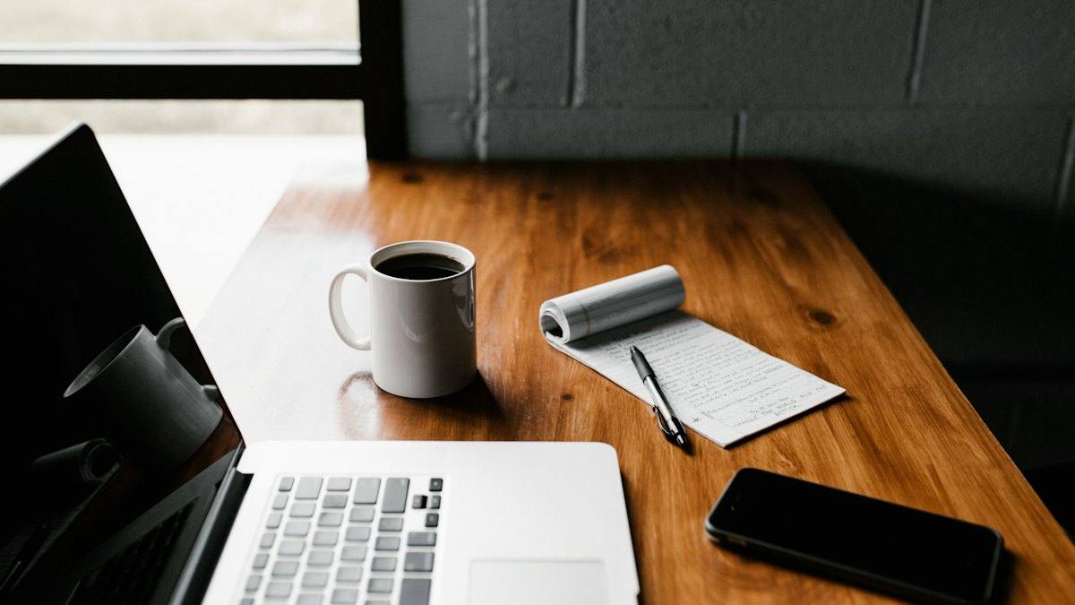 A laptop, coffee mug, notepad and phone on a wooden desk